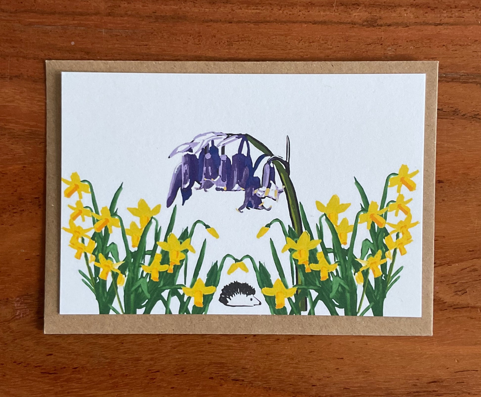 Close-up of wild notecard showing detailed hand-drawn bluebells, daffodils and hedgehog illustrations on premium 350gsm recycled cardstock texture.
